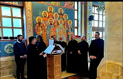 Sveštenici Žičke eparhije u prvom planu poju za pevnicom tokom Liturgije pređeosvećenih darova u Hramu Preobraženja Gospodnjeg na Zlatiboru. Kadar naglašava molitveni izraz i zajedništvo sveštenstva tokom bratske ispovesti i sastanka namesništava užičkog i račanskog. U pozadini se nazire unutrašnjost zlatiborskog hrama sa bogatim duborezom i freskopisom